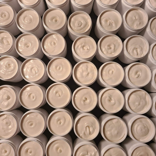 A close-up, top-down view of many open cosmetic jars filled with a creamy off-white product, arranged in a dense grid. The jars have white lids and light gray or beige outer containers, and the product itself has a warm cream tone.